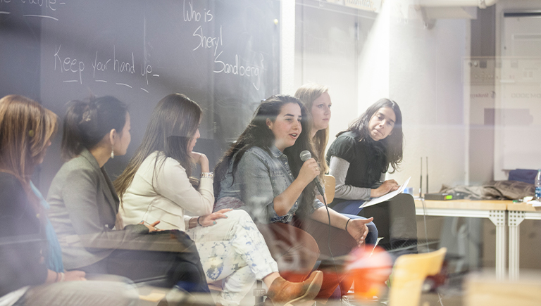 A group of female students looking at one student who is speaking into a microphone